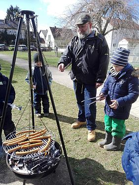 Wenn der Herd kalt bleibt, hilft ein Feuer: Hans Baldes grillt Würstchen für das Kindergarten-Mittagessen. Foto: Kath. Kita IN gGmbH/Faltermeier